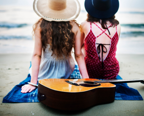 Musikinstrumente und Sportsachen Versicherung: Zwei junge Frauen sitzen am Strand mit dem Rücken zur Kamera und im Vordergrund liegt eine Gitarre auf dem Sand.