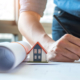 Eigenheim Finanzierung: Ein Mann mit eingerolltem Hausplan und einem kleinen Haus auf dem Tischt unterschreibt gerade seine Eigenheim Finanzierung.